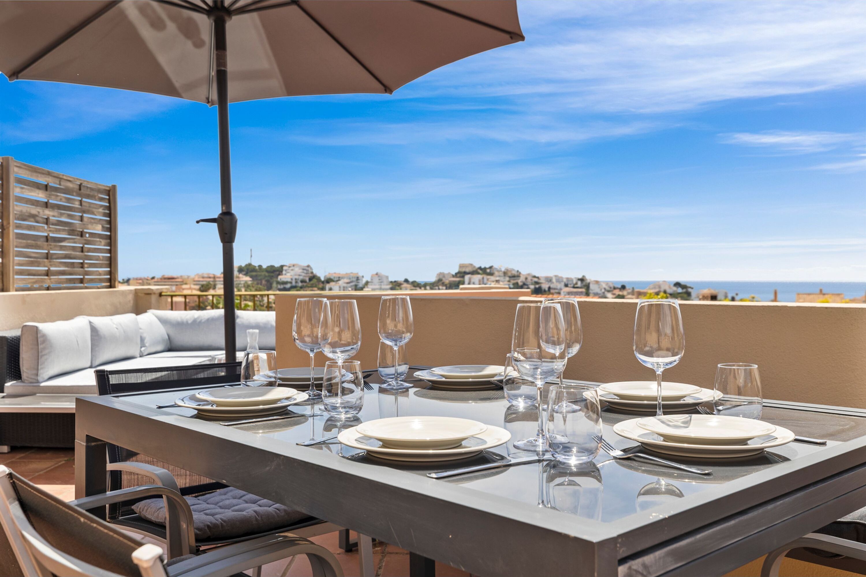 A dining table set with plates, glasses, and cutlery, under an umbrella, overlooking a scenic view.