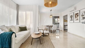 Una sala de estar moderna con un sofá blanco, una mesa de centro redonda de madera y un comedor con mesa y sillas.