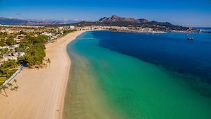 An aerial view of a coastal area with a sandy beach, clear blue waters, and a mountainous backdrop.