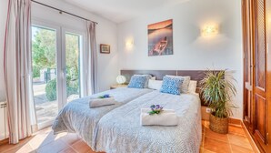 A double bed with a patterned bedspread, two pillows, a nightstand, a wall-mounted light, a framed picture, and a potted plant. - House Doris Hofmann Sa Ràpita with Pool (Sa Rapita)