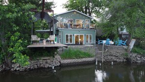A lakeside cabin with a deck, outdoor seating, and a stone wall.