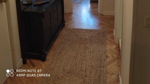 Un passadís amb una taula consola negra, un mirall i una làmpada penjant. Hi ha una porta a l'esquerra i una finestra amb cortines al fons.