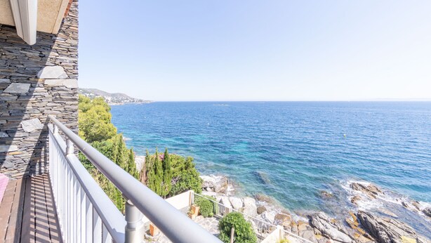 Ein Balkon mit Steinmauer und Geländer mit Blick auf eine Küstenlandschaft, das klare blaue Meer und eine felsige Küste zeigt.