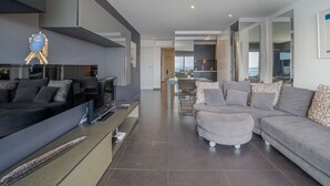 Ein modernes Wohnzimmer mit einer Couch, einem Couchtisch und einem Fernseher. Im Hintergrund befindet sich eine Küchenzeile.