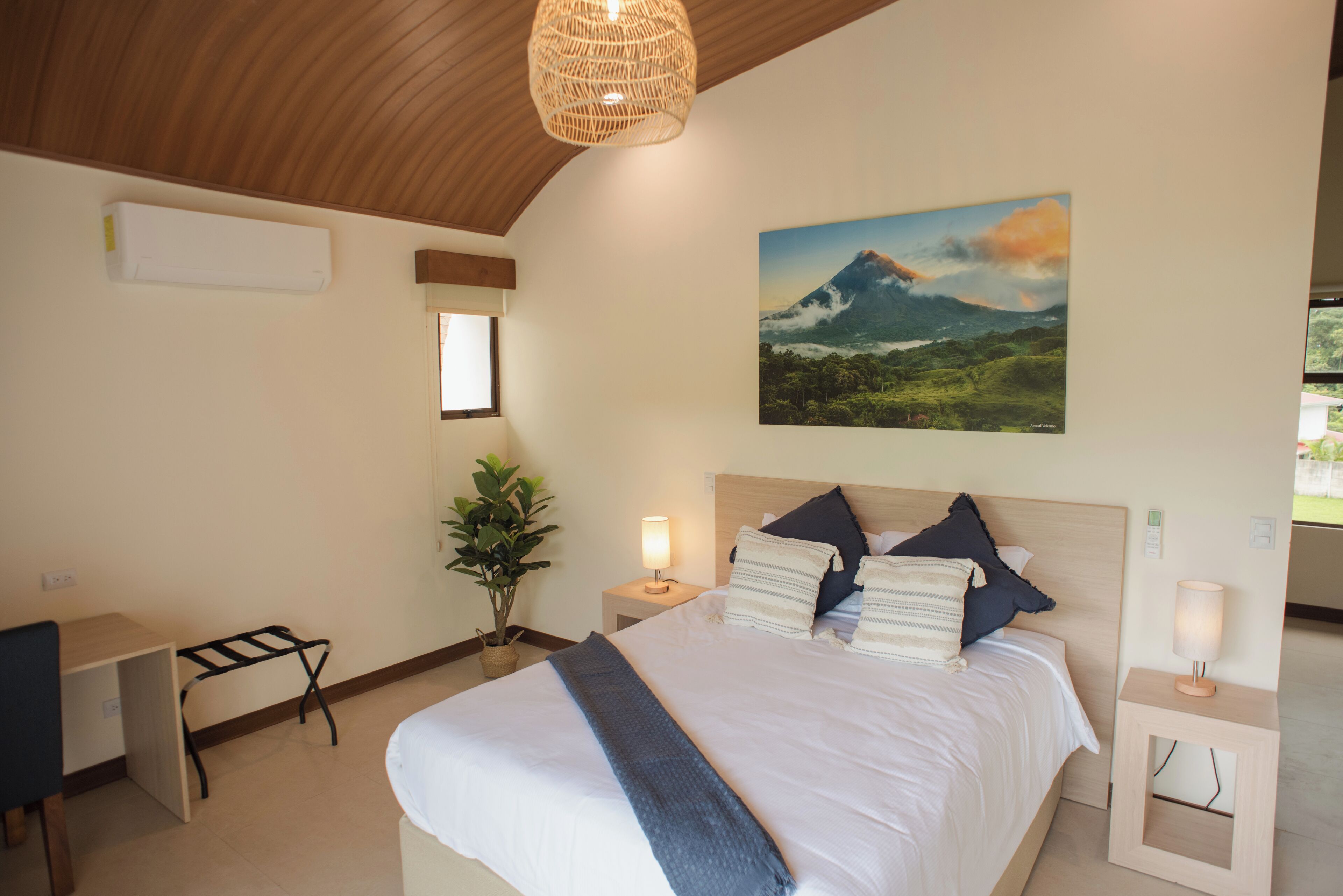 Ein Schlafzimmer mit Bett, Nachttischen, einem Schreibtisch, einer Topfpflanze und einem Gemälde einer Berglandschaft.
