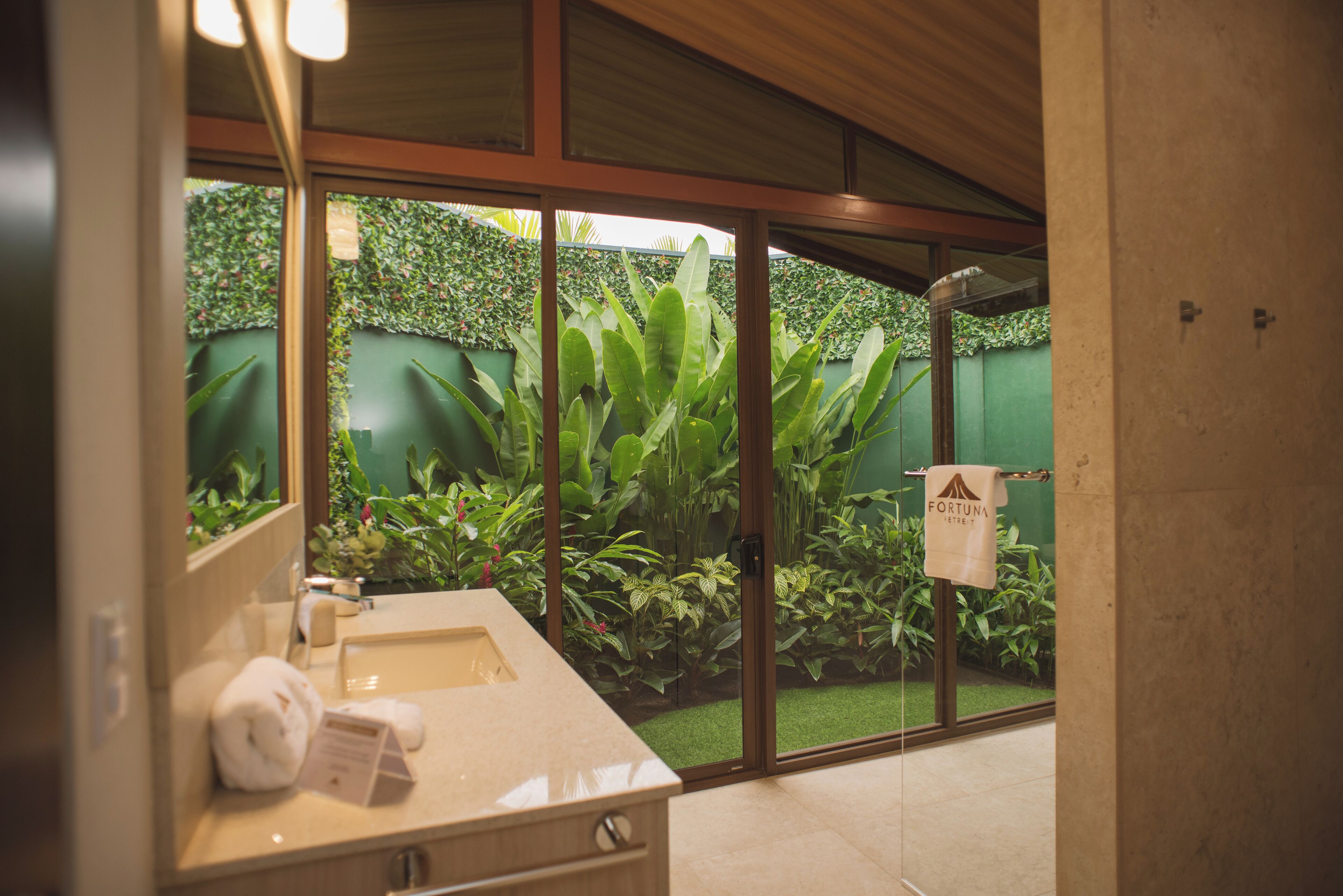 A bathroom with a large mirror, a sink, and a towel rack. There is a green wall and plants visible through the glass door.