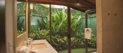 A bathroom with a large mirror, a sink, and a towel rack. There is a green wall and plants visible through the glass door.