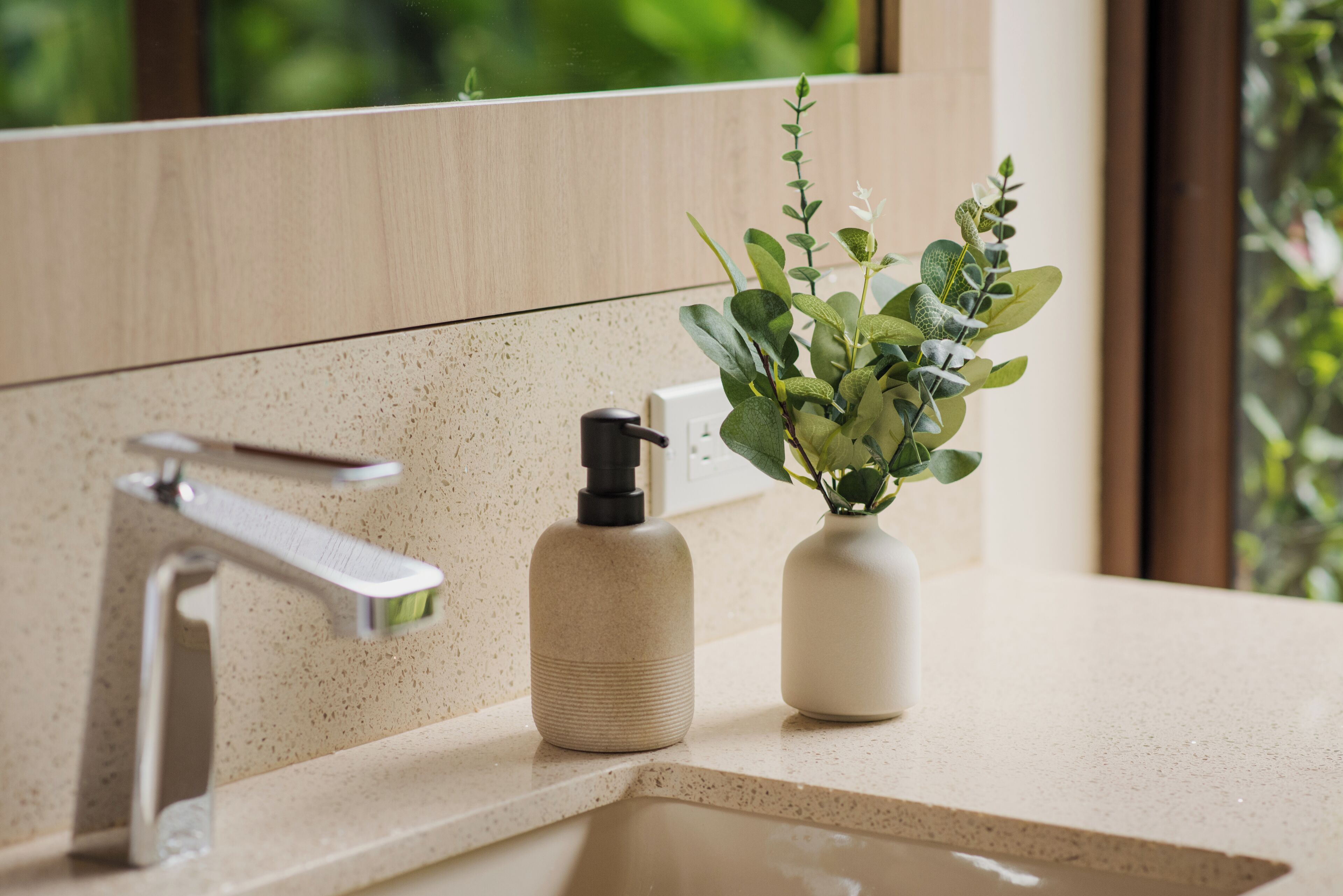 Ein Badezimmer mit einer beigen Ablagefläche, einem modernen Wasserhahn, einem Seifenspender und einem Blumentopf.