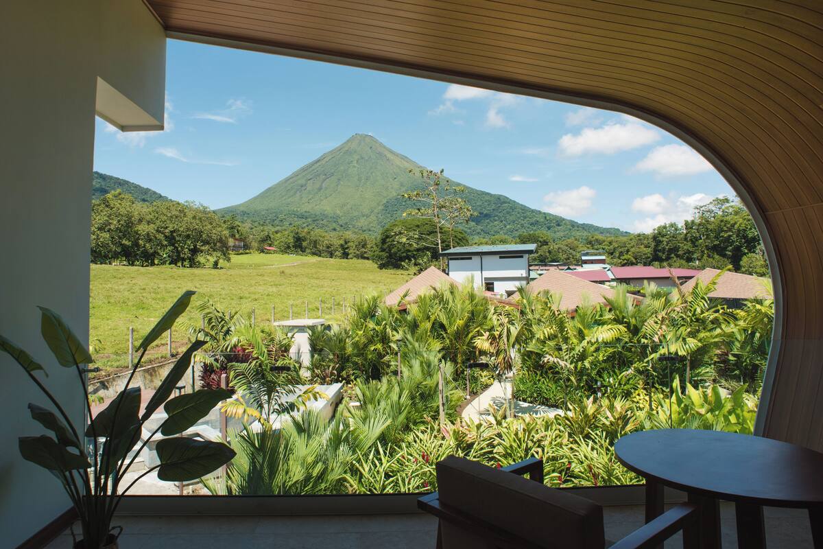 A view from a room with a large window overlooking a lush landscape, a mountain, and a few buildings.