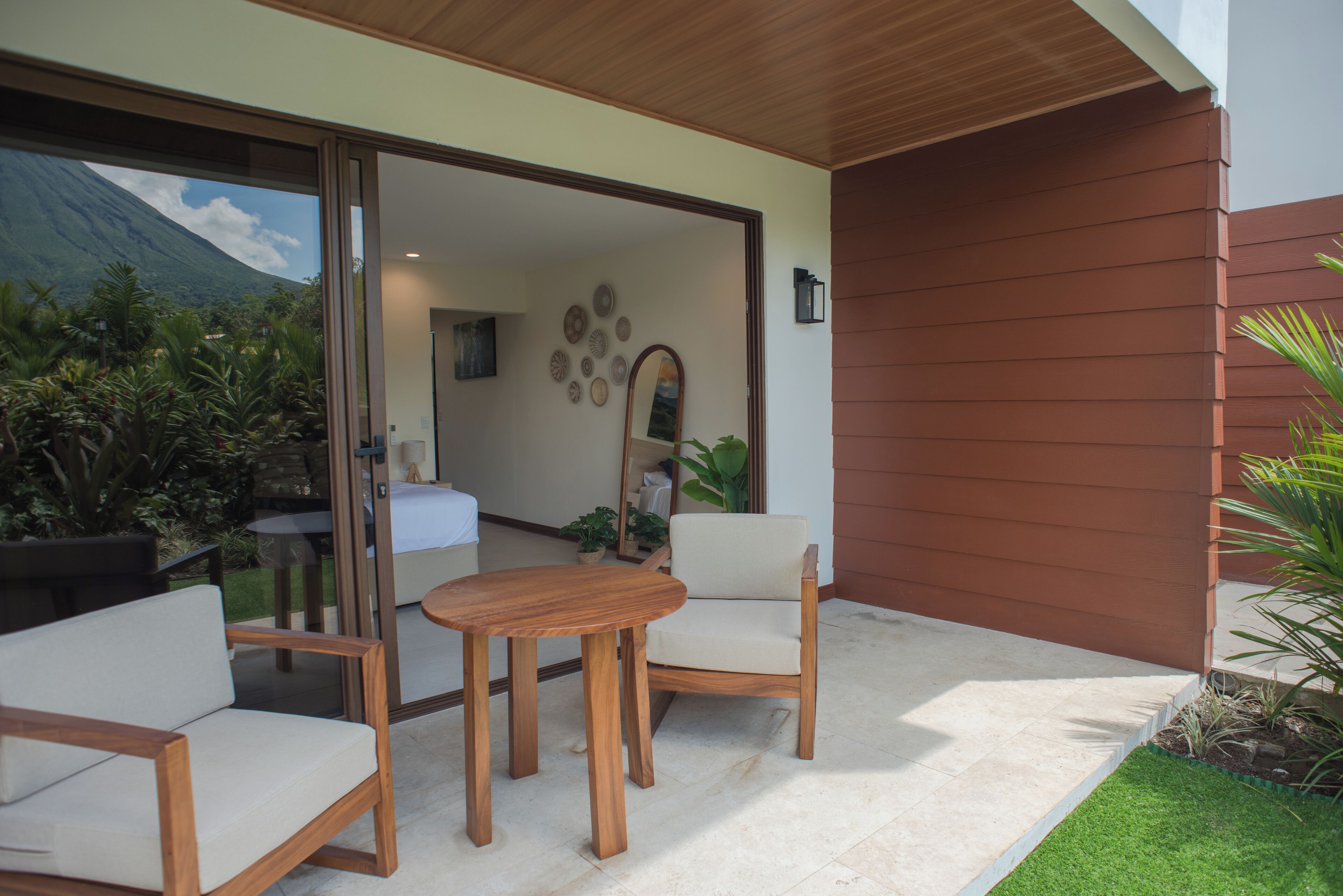 An outdoor patio with a wooden table and chairs, a glass door leading to a room with a bed and a wall decorated with circular mirrors.