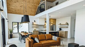 Un vaste espace de vie comprenant un canapé en cuir marron, une table basse en bois et une table à manger avec des chaises. On aperçoit une cuisine équipée d’appareils modernes et un balcon à l’arrière-plan.