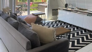 A modern living room with a sofa, a flat-screen TV mounted on the wall, a dining area with a table and chairs, and a view of greenery outside. - Modern A-FRAME cabin in the Ozark Mountains with amazing views! (West Fork)