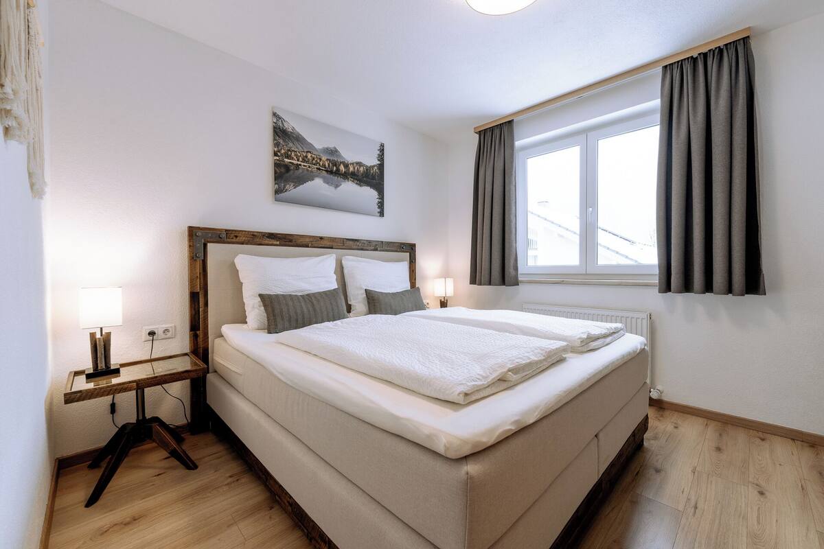 A bedroom with a large bed, a bedside table with a lamp, a window with curtains, and a painting on the wall.