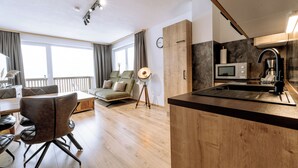 A modern living room with a sofa, dining table, and chairs. There is a kitchen area with a microwave and a coffee machine. The room has large windows and a balcony. - Apart Peak Ötztal  (Laengenfeld)