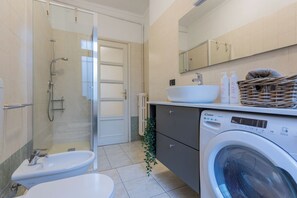 Un baño moderno con ducha envuelta en vidrio, lavamanos blanco, un bidé, una lavadora y un espejo sobre el lavamanos.