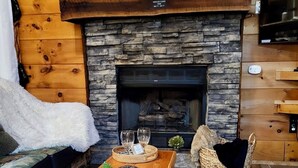 Ein gemütliches Blockhaus-Interieur mit Kamin aus Stein, einem hölzernen Schild darüber und einem rustikalen Couchtisch in der Mitte.