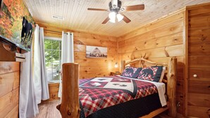 Ein Schlafzimmer einer Holzhütte mit einem Bett, einem Deckenventilator, einem Fenster mit Vorhängen und einem Bärenbild an der Wand.