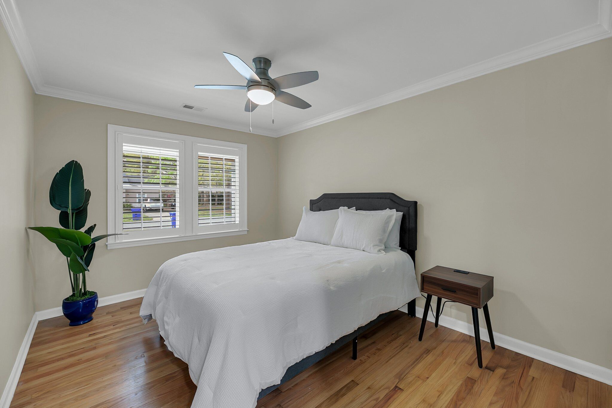 A bedroom with a bed, a ceiling fan, a window with blinds, and a potted plant.