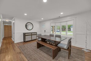 Un comedor con una mesa de madera, bancos y sillas. Hay un ventanal con persianas, una puerta y un espejo en la pared.