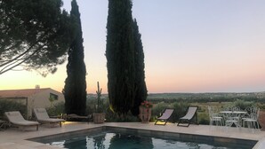 A rectangular swimming pool with lounge chairs and a table, surrounded by tall cypress trees and a view of the countryside. - Maison (Maraussan)