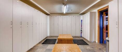 A locker room with multiple lockers, a wooden bench, and a trash can.
