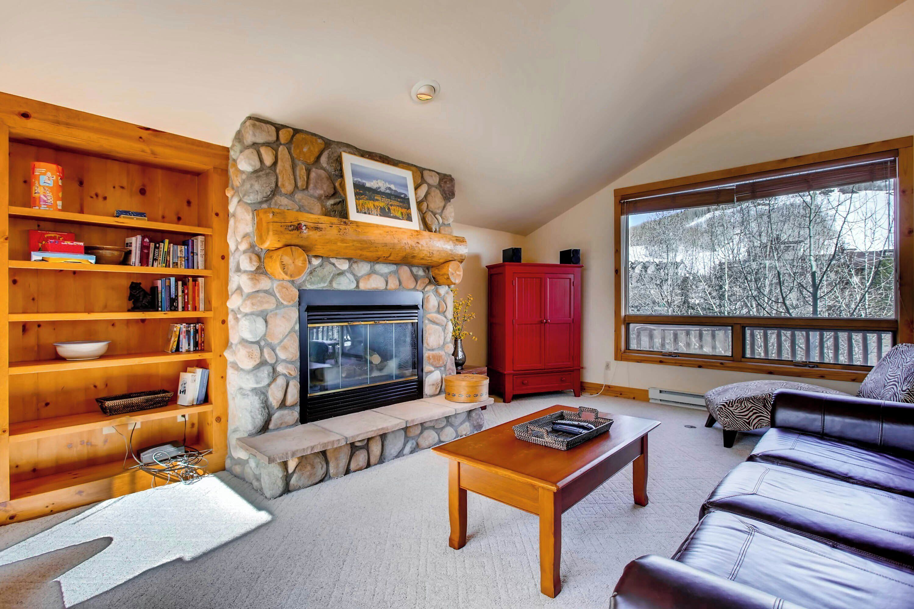 A cozy living room with a stone fireplace, wooden shelves, a red cabinet, a leather sofa, and a large window with a view of trees.