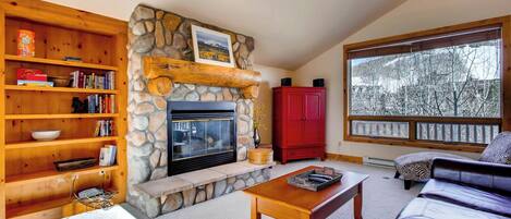 A cozy living room with a stone fireplace, wooden shelves, a red cabinet, a leather sofa, and a large window with a view of trees.