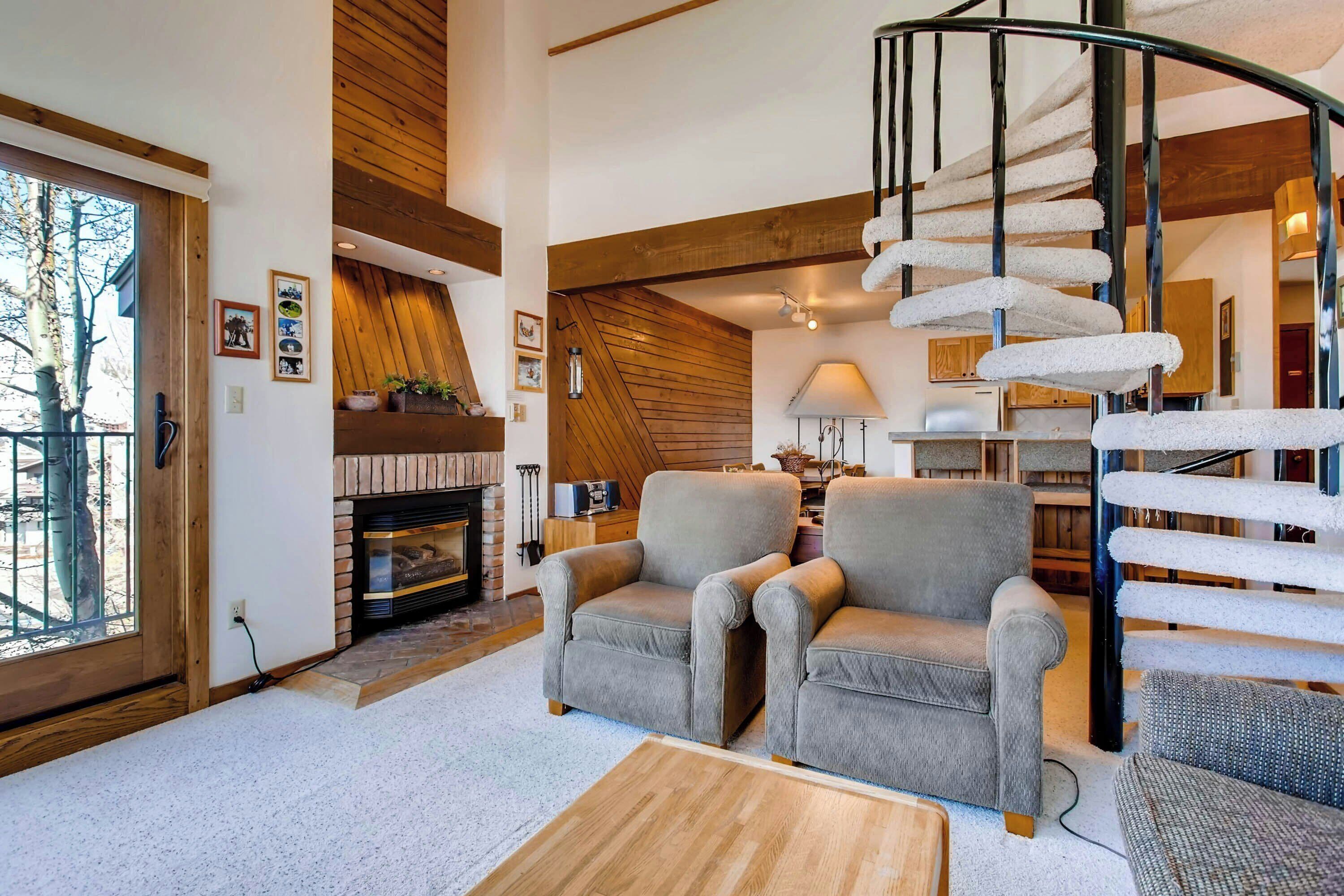 A modern living room with a fireplace, two armchairs, a wooden coffee table, and a spiral staircase.