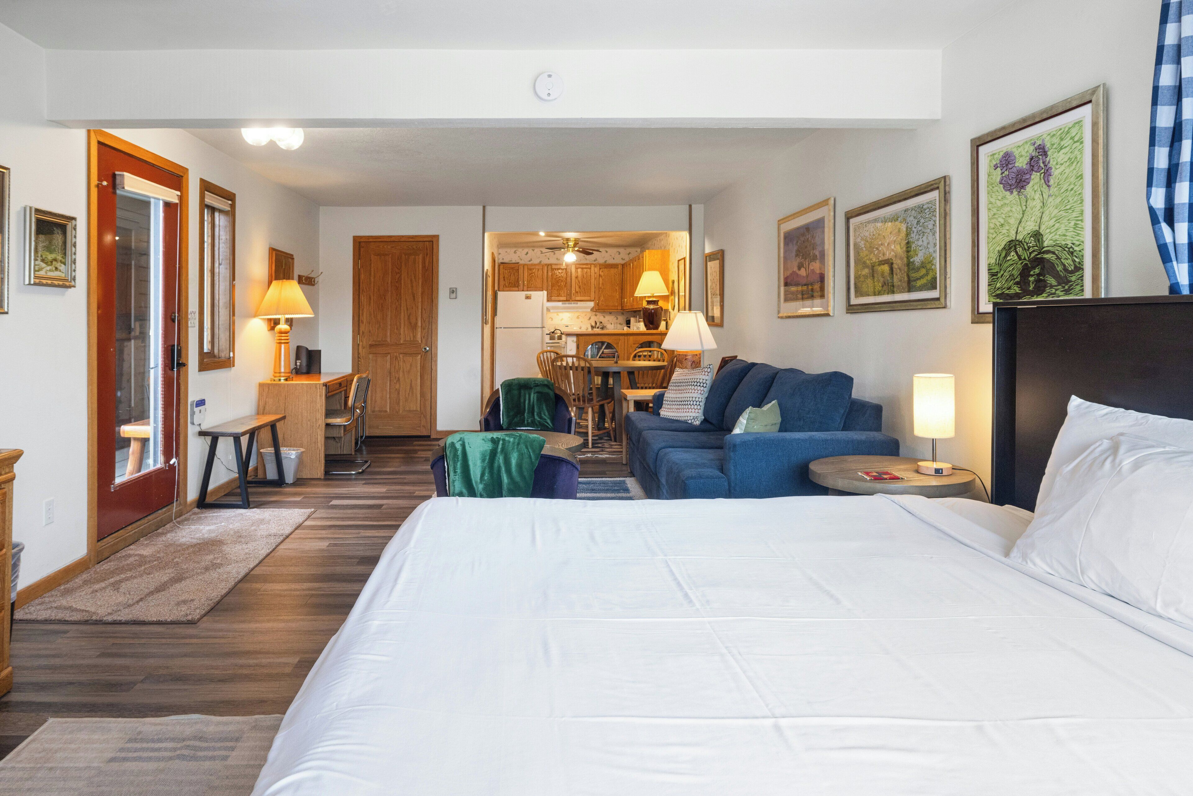 A hotel room with a large bed, a sofa, a dining table, and a kitchen area visible through an open door.