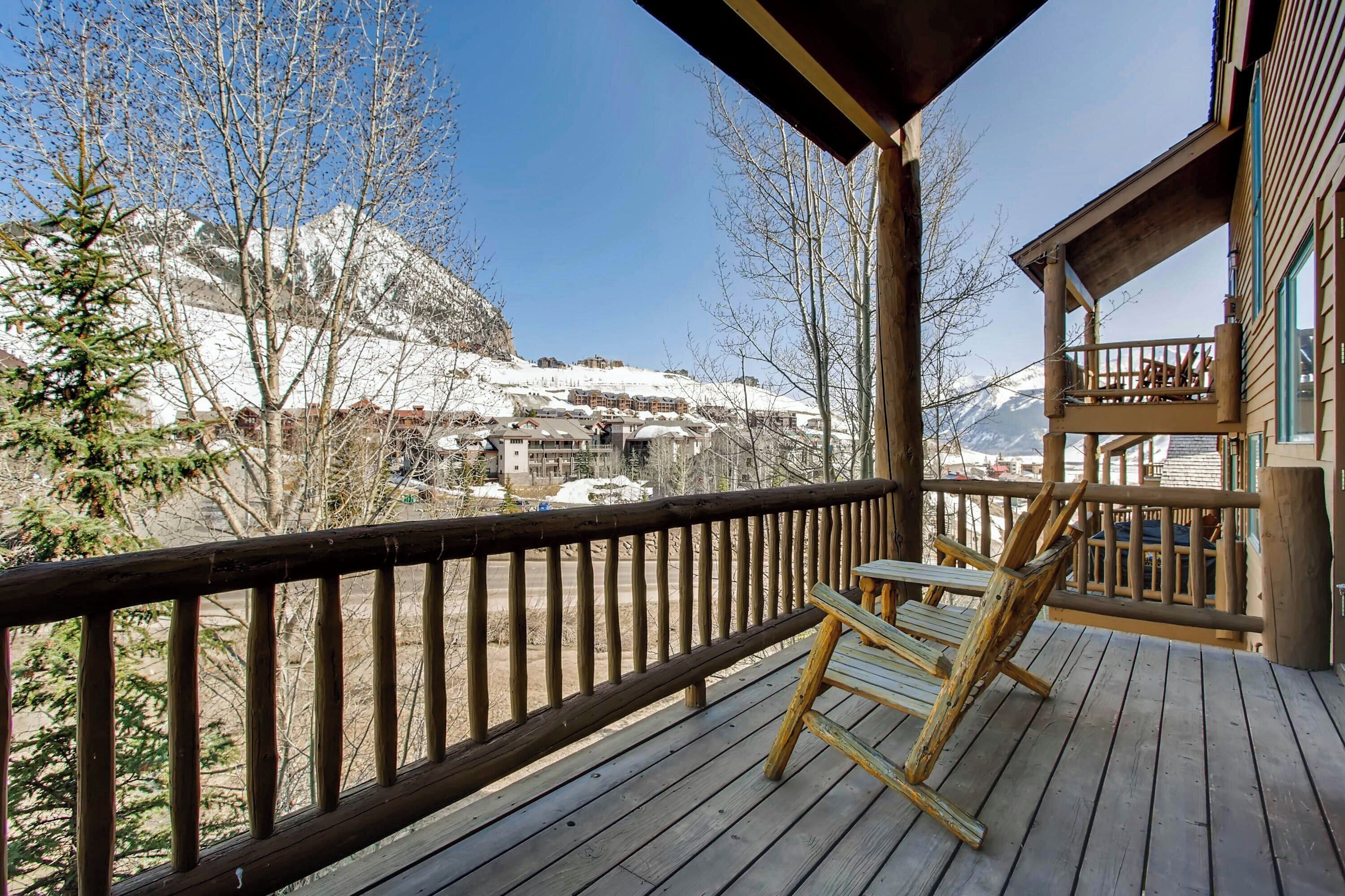 Une terrasse en bois avec une chaise et une table, d’où l’on peut admirer un paysage montagneux enneigé, avec des bâtiments visibles au loin.