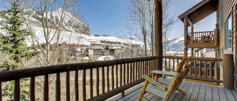 A wooden deck with a chair and a table, overlooking a snowy mountain landscape with buildings in the background.