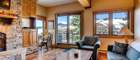 A cozy living room with a stone fireplace, wooden beams, a large window with a mountain view, a sofa, and a coffee table.