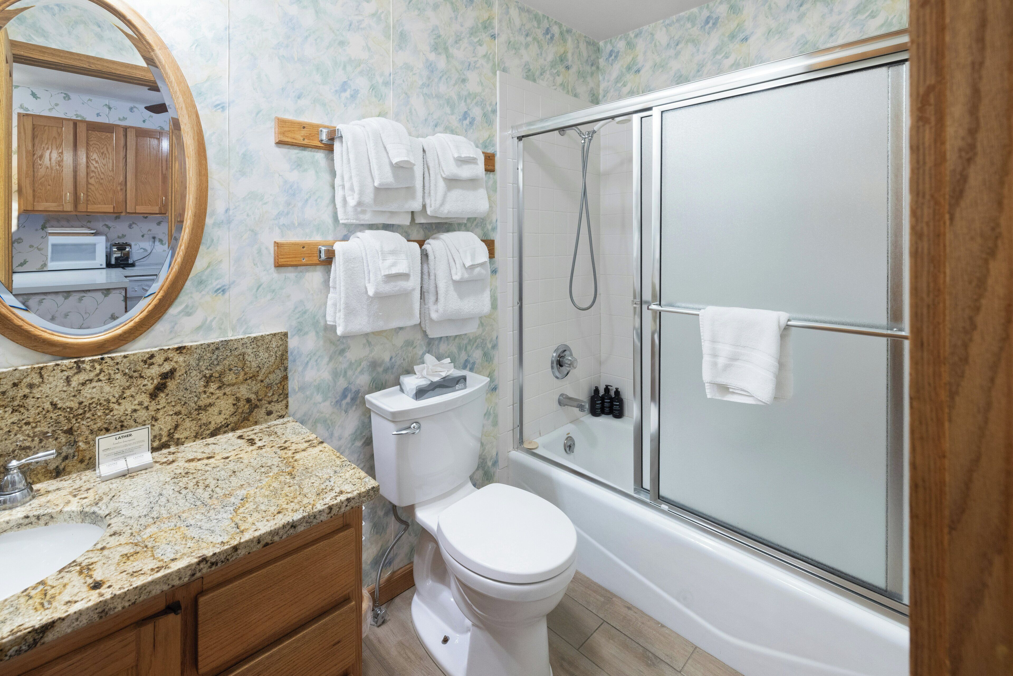 Baño con inodoro, lavamanos y ducha. Hay un espejo sobre el lavamanos y un perchero con toallas.