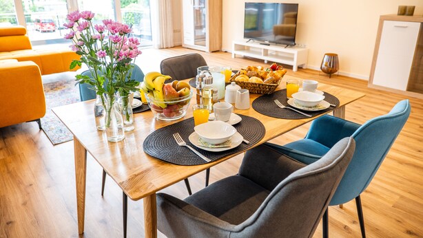 Ein Frühstückstisch mit Holz, Stühlen, einer Obstschale, einem Blumenstrauß und einem Fernseher im Hintergrund.