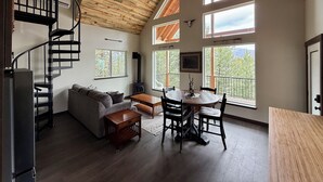Un vaste espace de vie doté d’un plafond en bois, d’un escalier en colimaçon, d’une table à manger avec des chaises, d’un canapé et de grandes fenêtres offrant une vue sur les arbres et les montagnes.