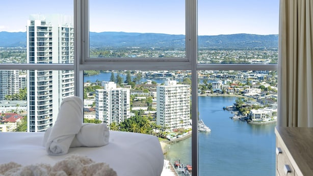 A hotel room with a large window offering a view of a cityscape and a body of water. - Two-Bedroom Apartment with Water View (SURFERS PARADISE)