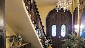 A grand staircase with a dark wooden finish and intricate balusters, leading to a large, ornate door with glass panels. A chandelier hangs from the ceiling, and the floor is tiled in a black and white checkered pattern. - 1873 Victorian Rose B & B (Port Washington)