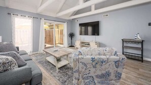 A spacious living room with a grey sofa, a floral armchair, a wooden coffee table, and a flat-screen TV mounted on the wall.