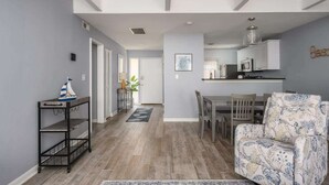A modern living room with a sofa, a dining table with chairs, and a kitchen area with cabinets and a hanging light fixture.