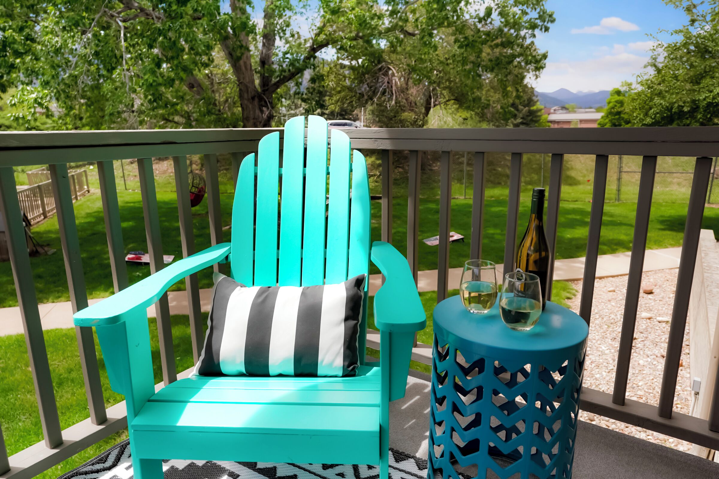 Een turquoise Adirondack-stoel met een gestreept kussen op een balkon met uitzicht op een gazon en bomen.