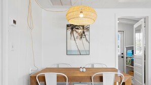 A dining area with a wooden table and white chairs, a wall-mounted picture of palm trees, and a hanging wicker light fixture.