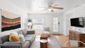 A modern living room with a grey sofa, a wooden coffee table, a ceiling fan, a TV, and a large wall tapestry.