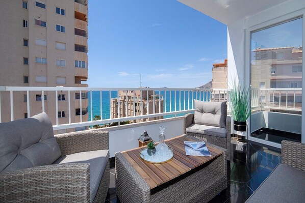 A balcony with wicker furniture, a table with a bottle and a menu, and a view of the sea and buildings. - Apartment 'La Reina' with Sea View, Wi-Fi and Air Conditioning (Calpe)