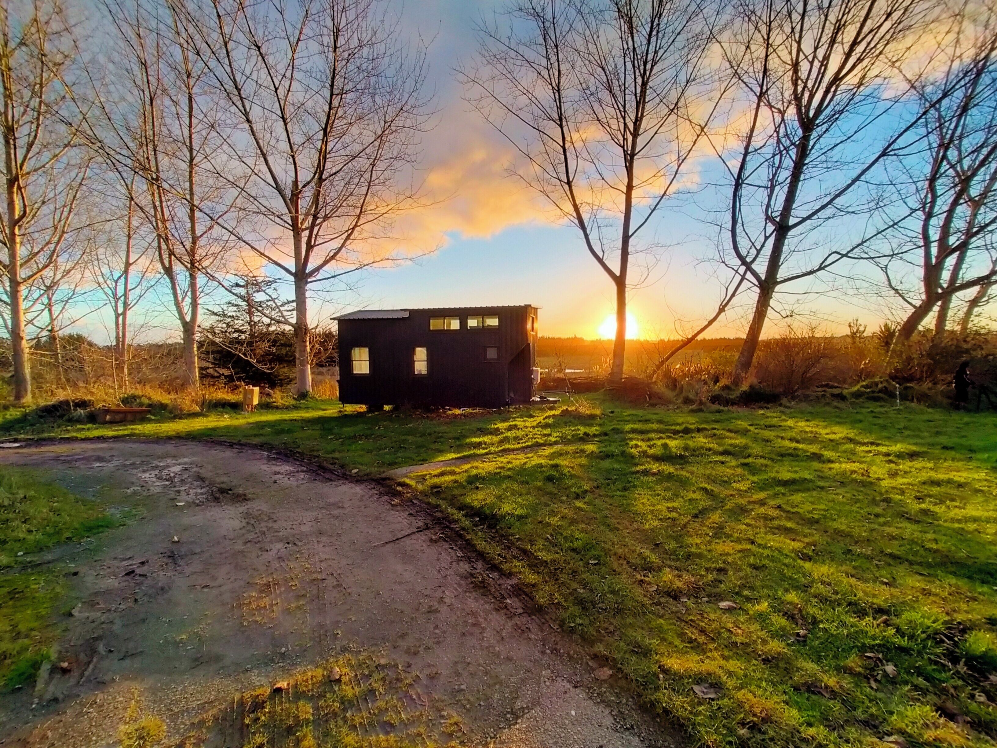 A rustic cabin with a dirt path leading to it, surrounded by trees and a sunset in the background.
