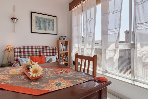 A cozy living room with a sofa, a wooden table with a floral table runner, a bookshelf, and a window with sheer curtains.