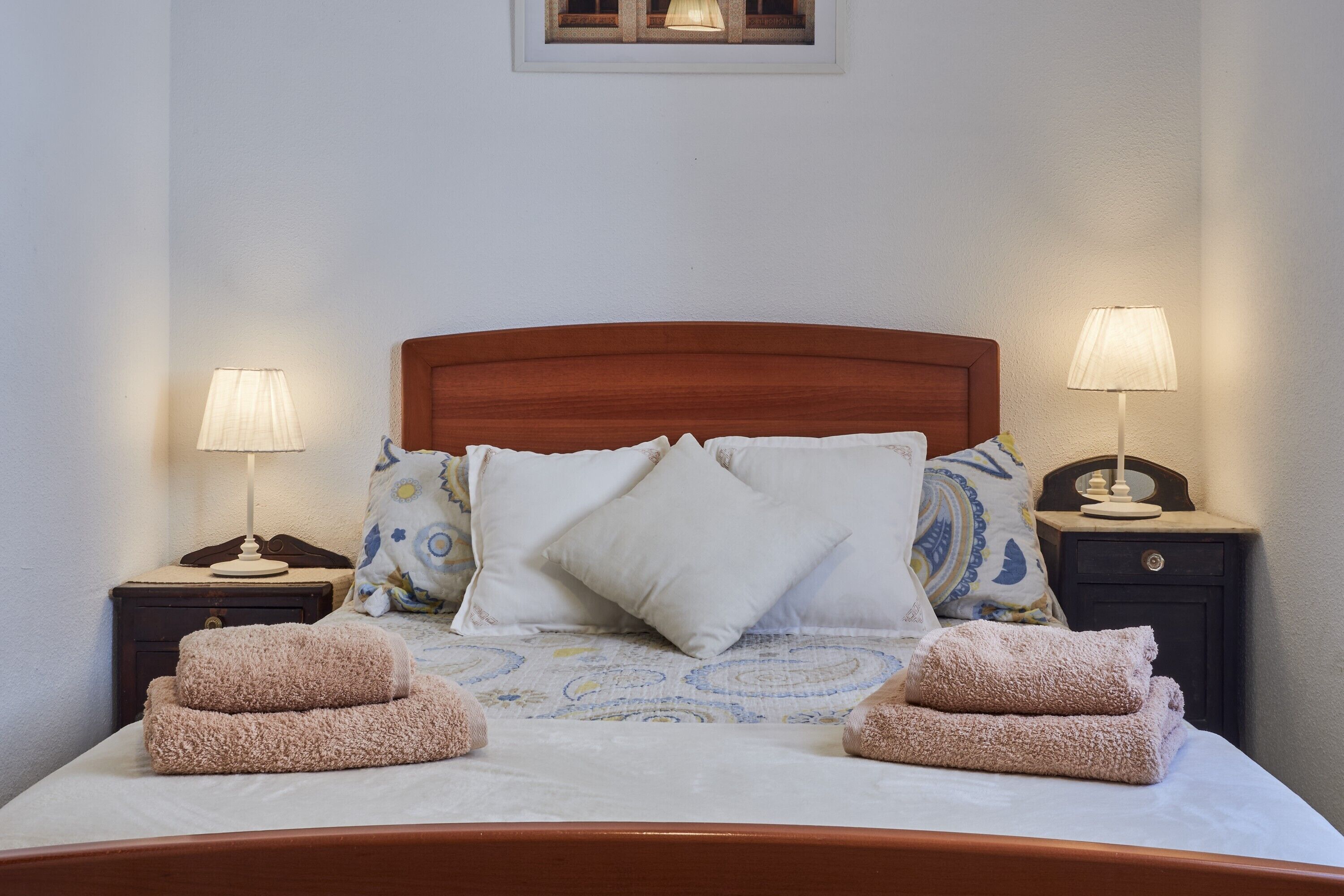 Una cama bien tendida con cabecera de madera, dos mesitas de noche con lámparas y una ventana arriba.