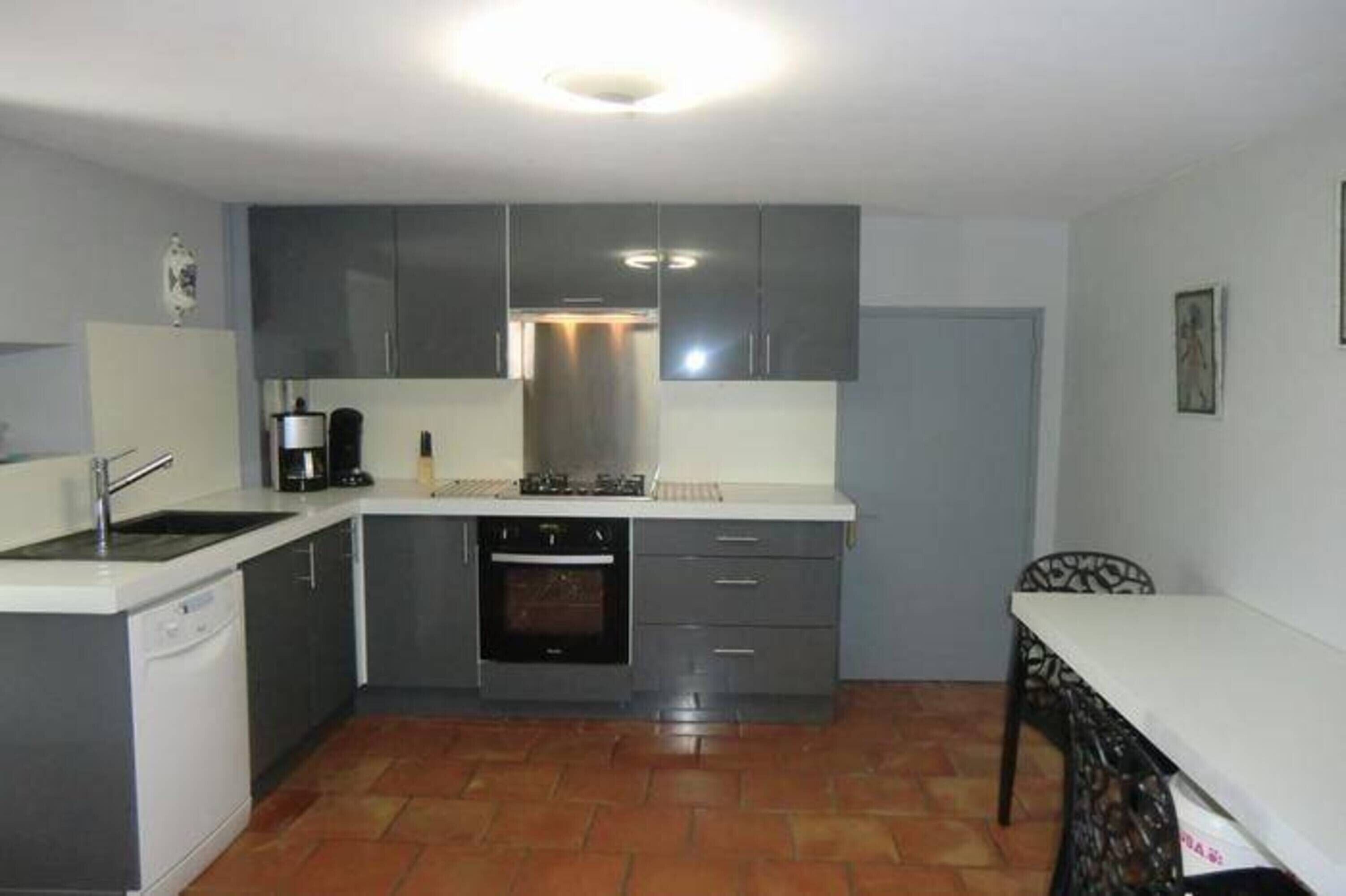 A compact kitchen with grey cabinets, a white countertop, a built-in oven, and a dining area with a table and chair.