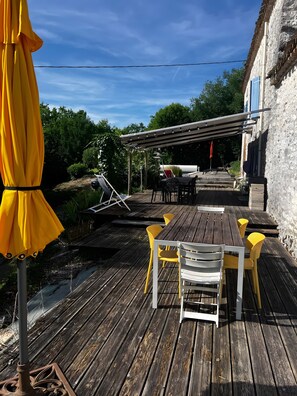 A wooden deck with a table and chairs, a pergola with lights, and a building with blue shutters. - La Boissière – Peaceful White Stone Retreat with Pool and Air Conditioning (Fauroux)
