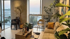 A modern living room with a sofa, coffee table, and chairs, large windows with a view of buildings and the sea.