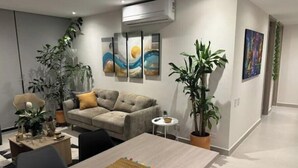 A modern living room with a sofa, a coffee table, and a dining table. There are plants, artwork on the wall, and an air conditioning unit.